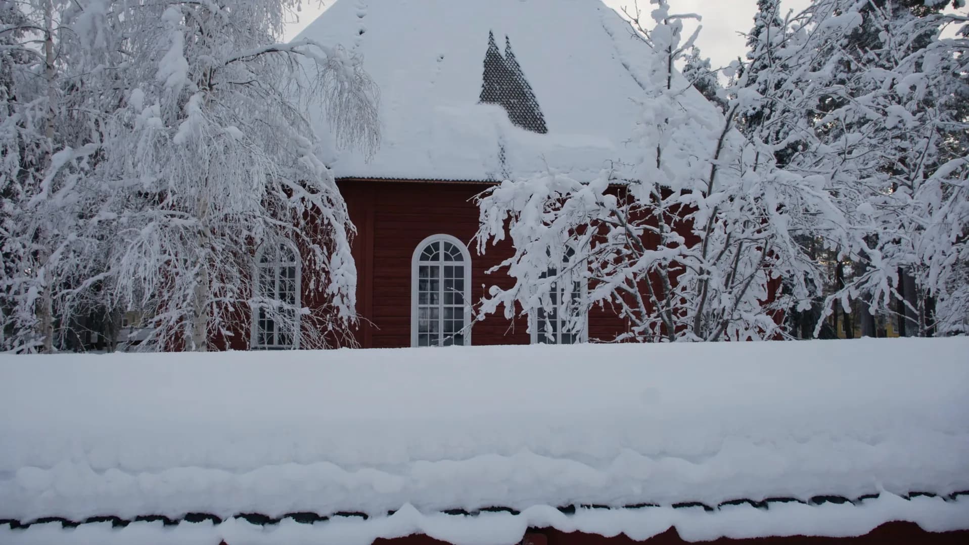 Red timber walls and an arched white window of the Old Church in Jokkmokk, half hidden by snow-laden birches behind a snow-covered wall