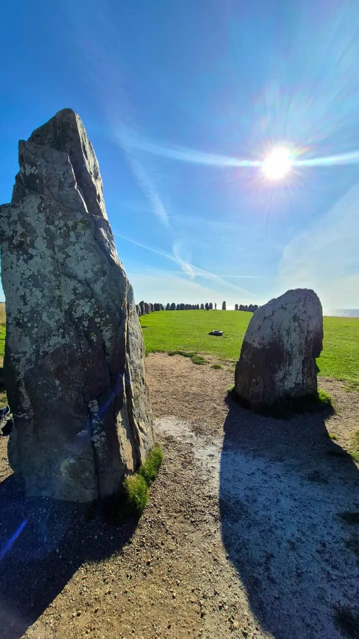 Two of the standing stones of Ales stenar casting long shadows across a gravel path under a low, bright sun, the rest of the stone ship and the sea beyond, a frame from a short film