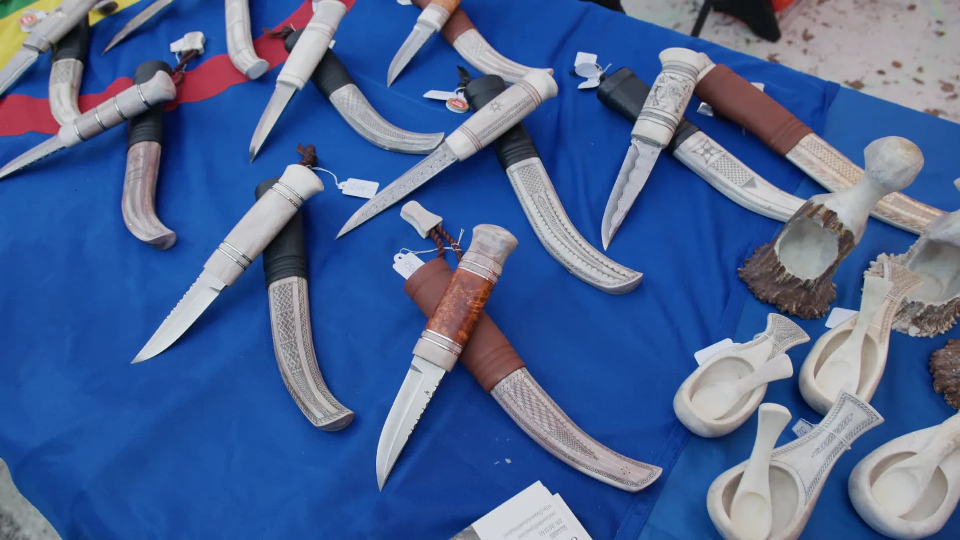 Sámi knives with reindeer antler handles and engraved antler sheaths on a blue cloth beside small antler cups, on a stall at the Jokkmokk winter market