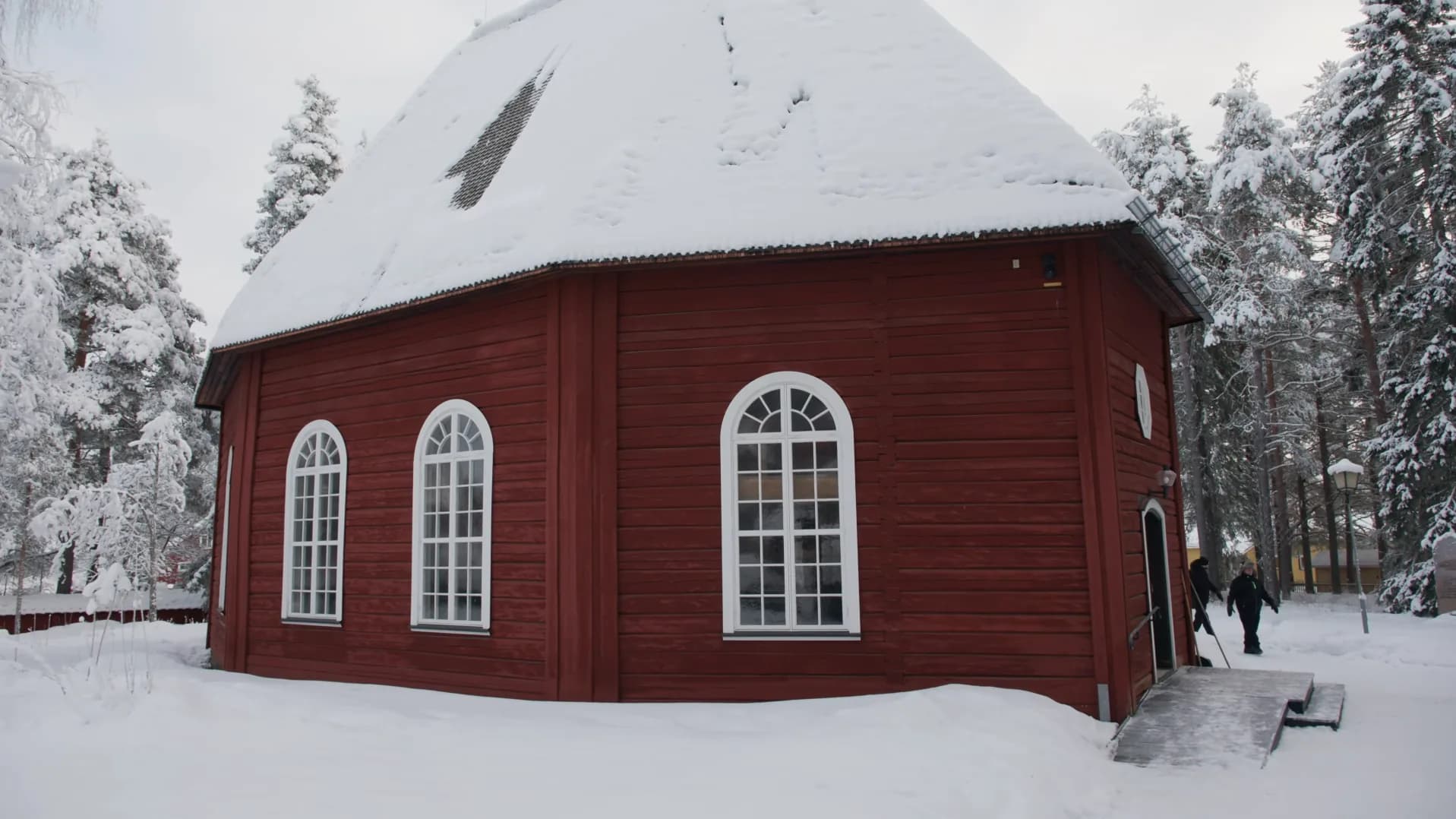 Red eight-sided timber church with arched white windows and a snow-covered roof among frosted pines, the Old Church in Jokkmokk in winter