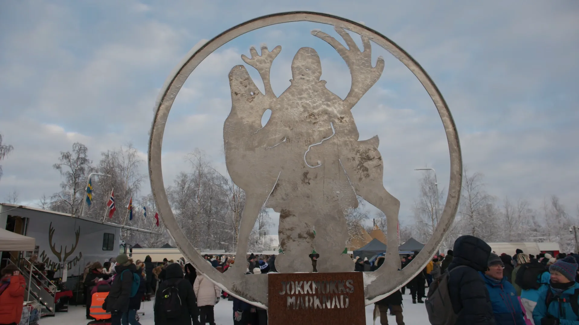 Steel sculpture of a figure and a reindeer inside a ring on a plinth lettered Jokkmokks Marknad, with market stalls, flags and crowds beyond under a winter sky