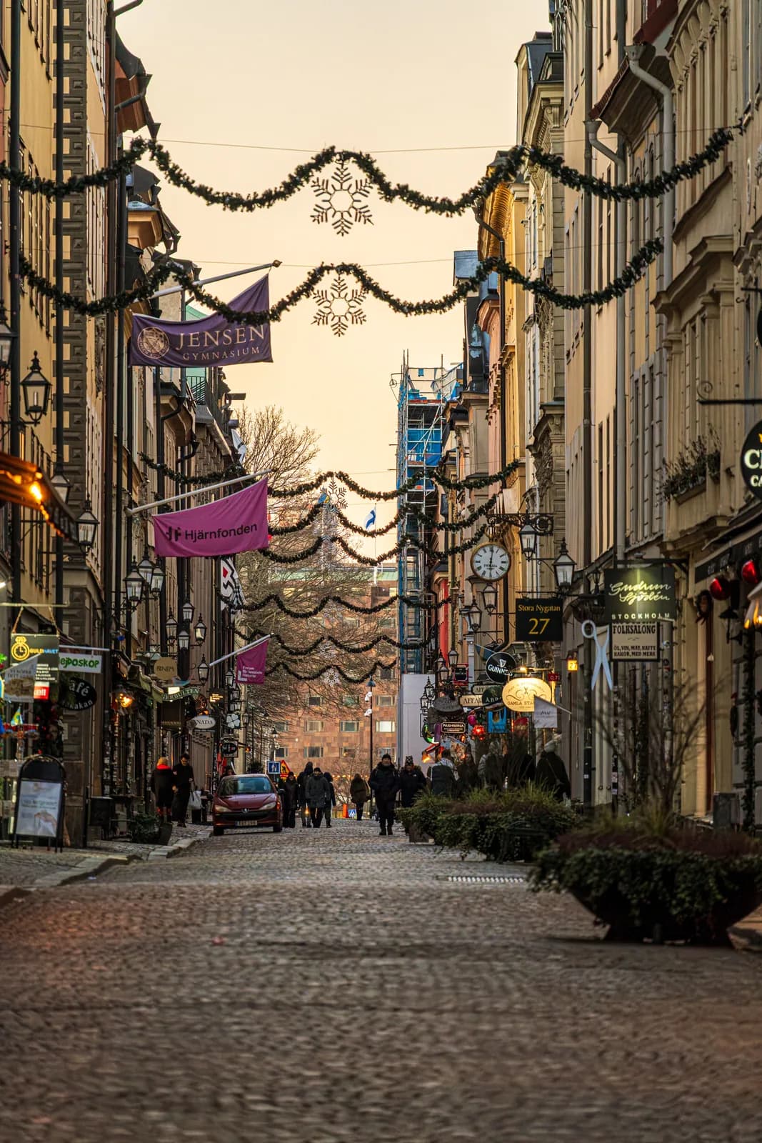 Christmas garlands and snowflake lights strung across Stora Nygatan in Gamla stan, Stockholm, with shops and people walking on the cobbles at dusk