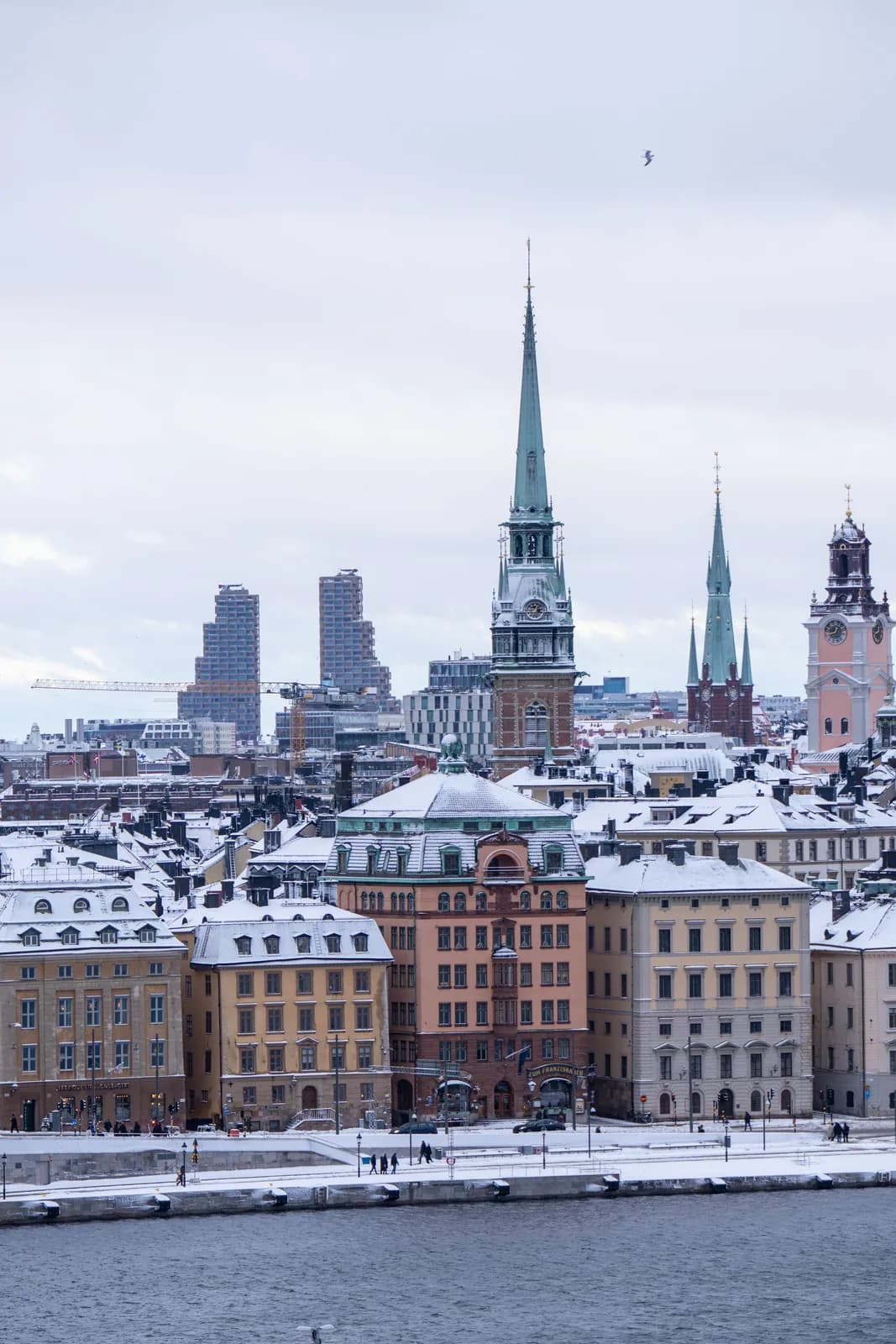 Snow on the roofs of Gamla stan in Stockholm, seen across the water, with the green spire of the German Church and other church towers under a grey winter sky