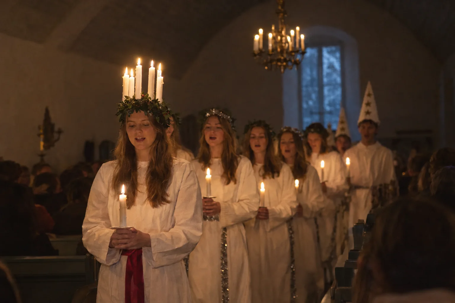 Lucia in a white gown and red sash, wearing a crown of lit candles, leads a candlelit procession of singers in white and star boys in tall pointed hats down the aisle of a dim whitewashed church