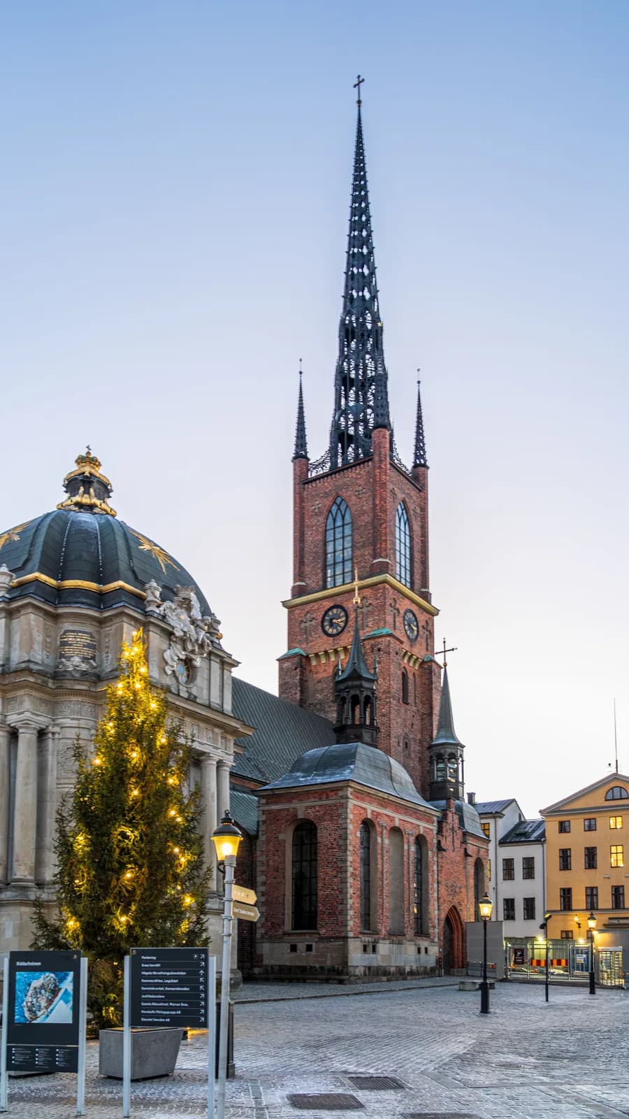 Riddarholmen Church in Stockholm at dusk in winter, its brick tower and iron spire beside a lit Christmas tree on the cobbled square