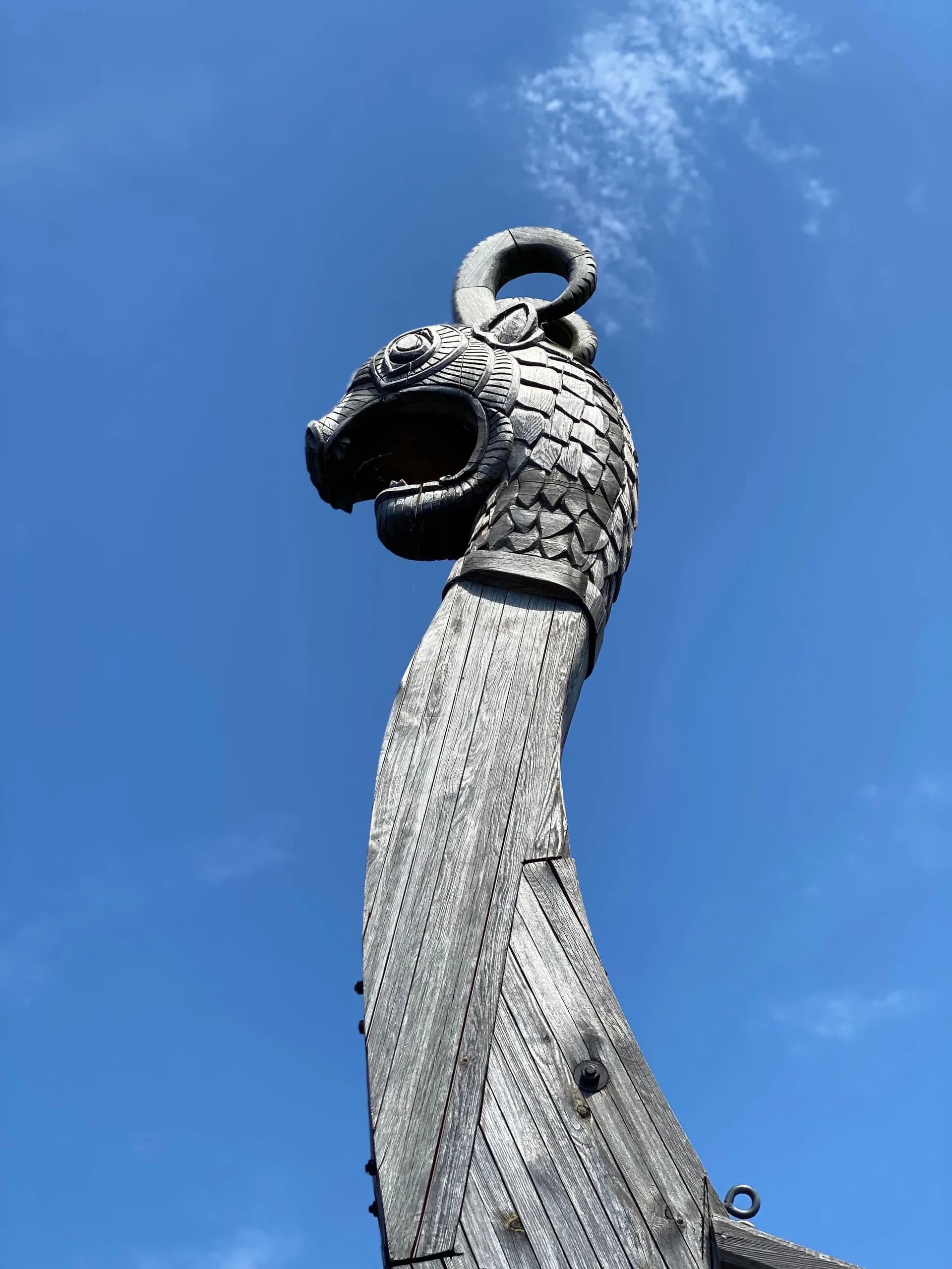 Carved wooden dragon head on the tall prow of a replica Viking ship, seen from below against a blue sky