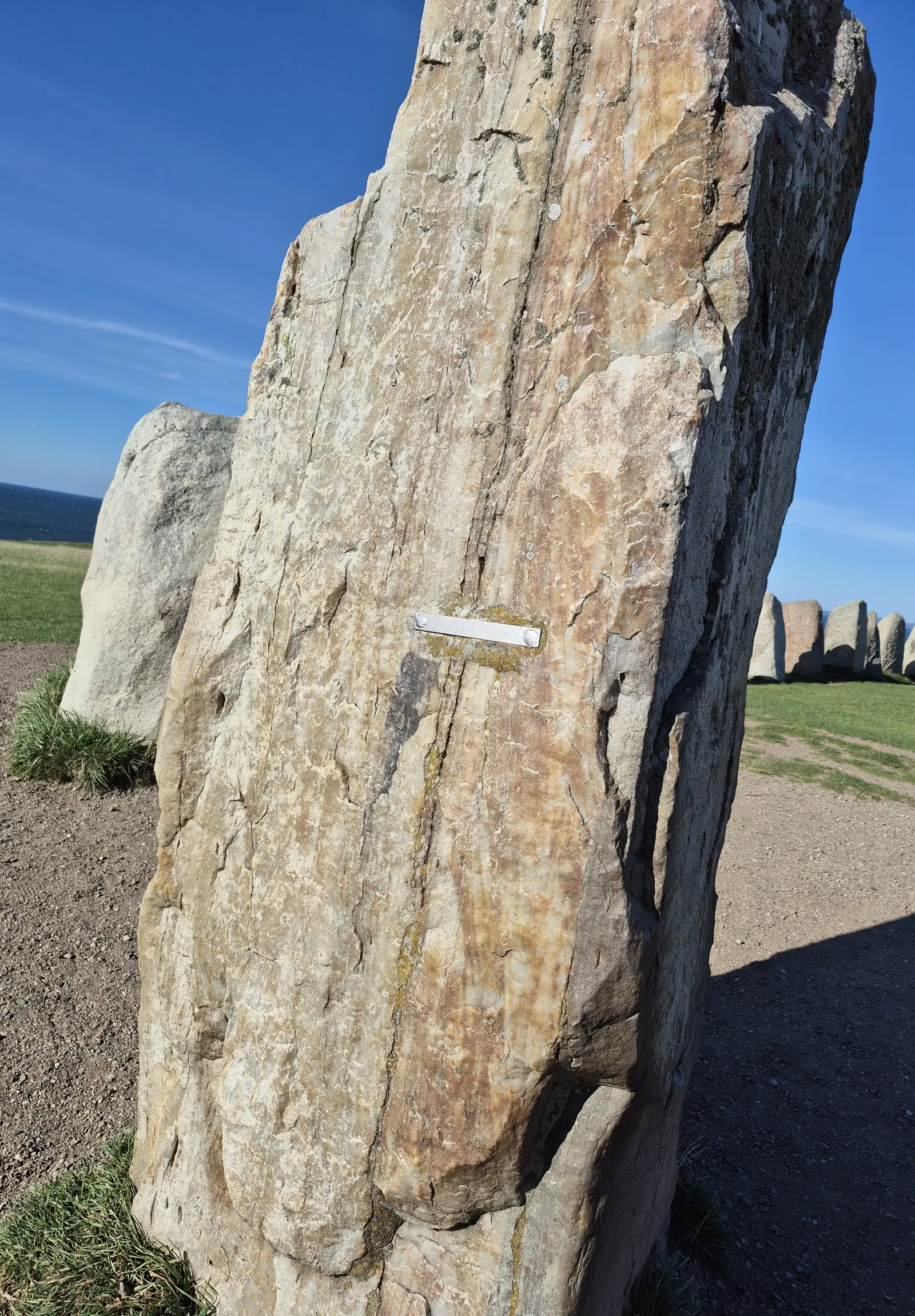 Close view of one tall weathered standing stone at Ales stenar, streaked pink and grey, with more stones of the ship and the sea behind