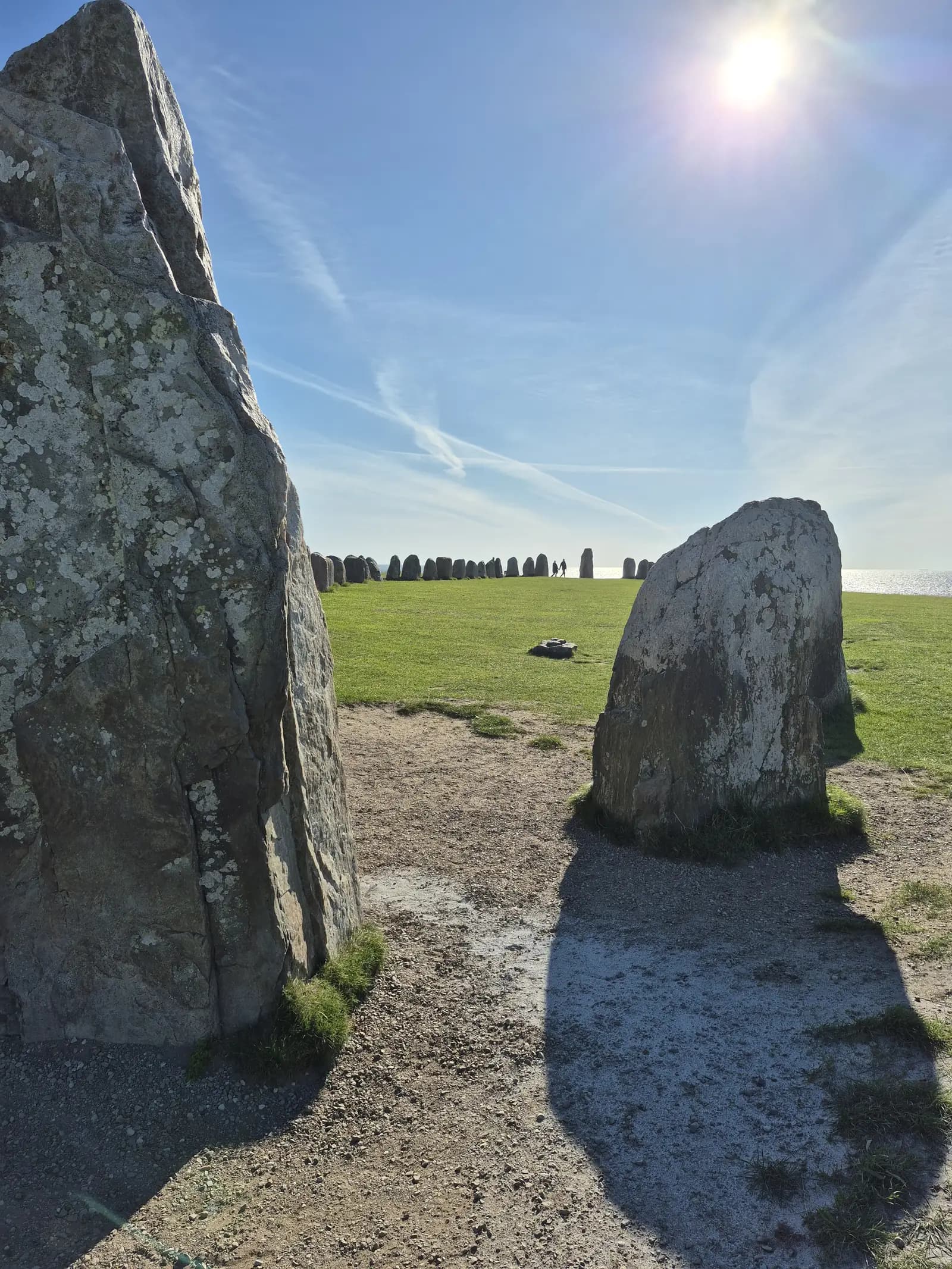 Two standing stones framing the length of the Ales stenar stone ship on its grassy headland, the sea beyond and a low sun in a blue sky