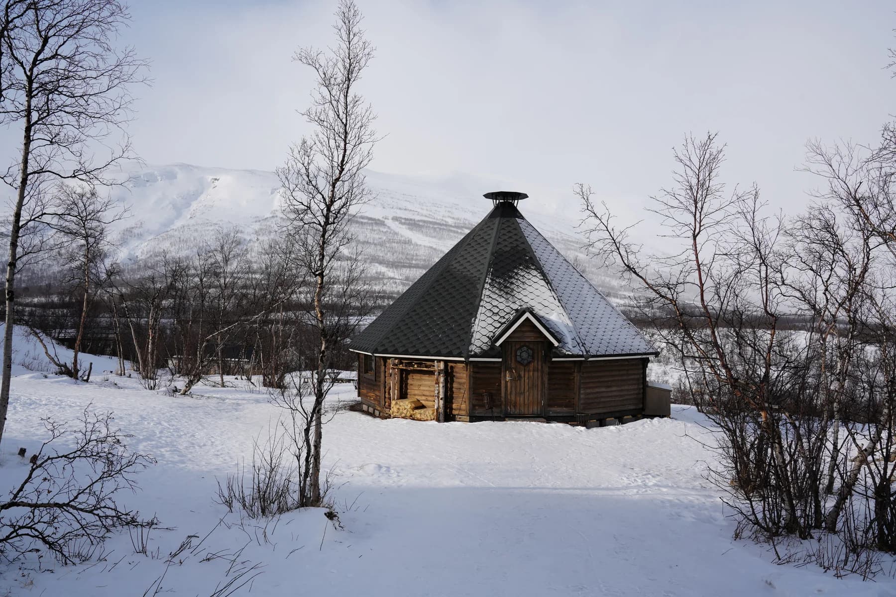 Octagonal wooden hut with a dark shingled roof in deep snow among bare birches, with snowy mountains behind