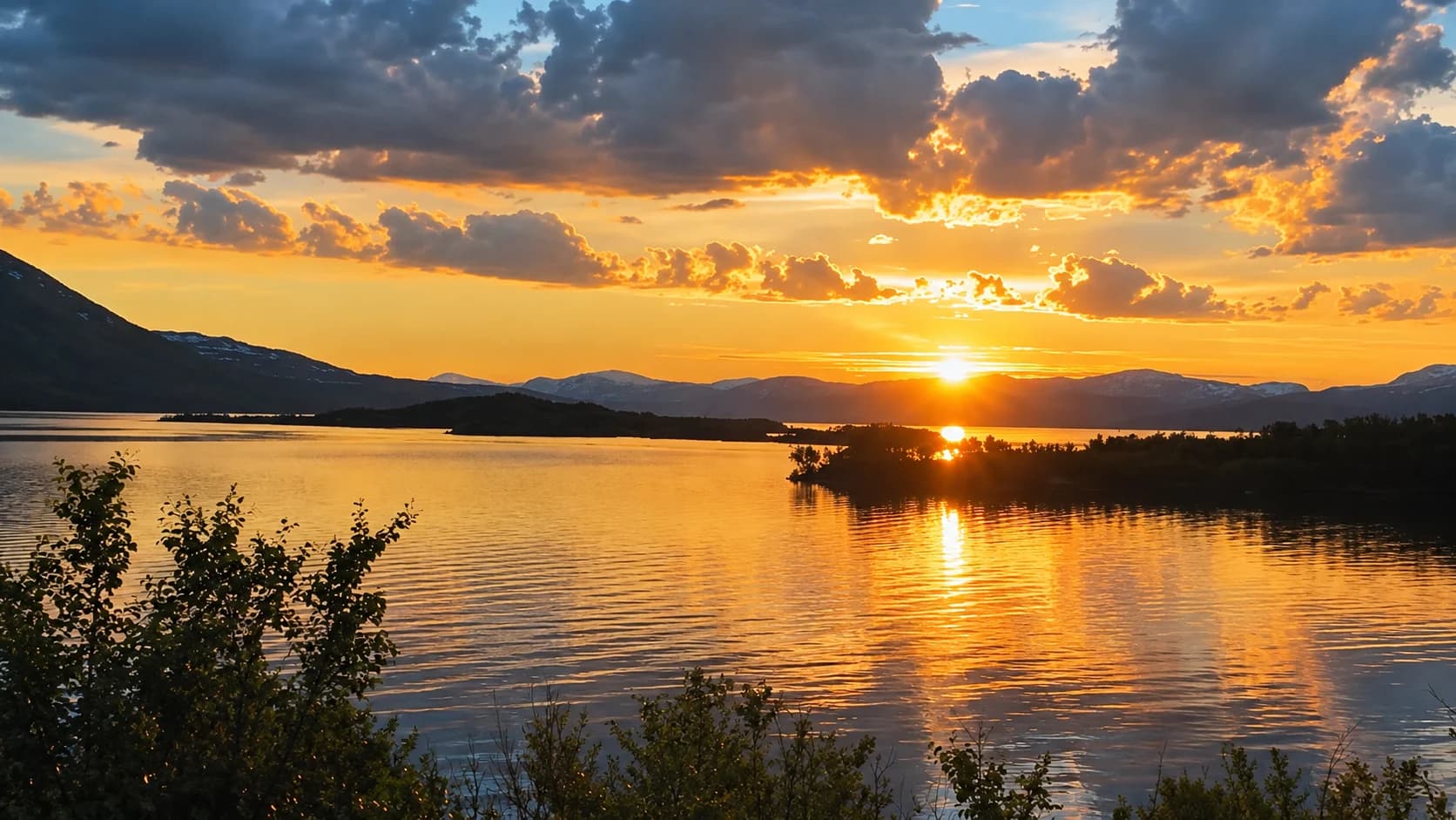 Low golden sun above the mountains, reflected across the lake at Abisko, with birch scrub in the foreground