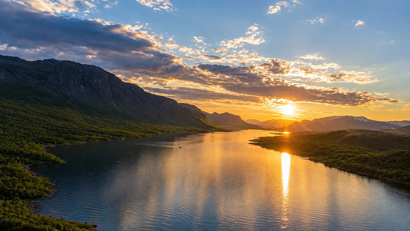 Low sun over a long mountain lake between birch-covered slopes, its light reflected down the water, with a small boat on the lake