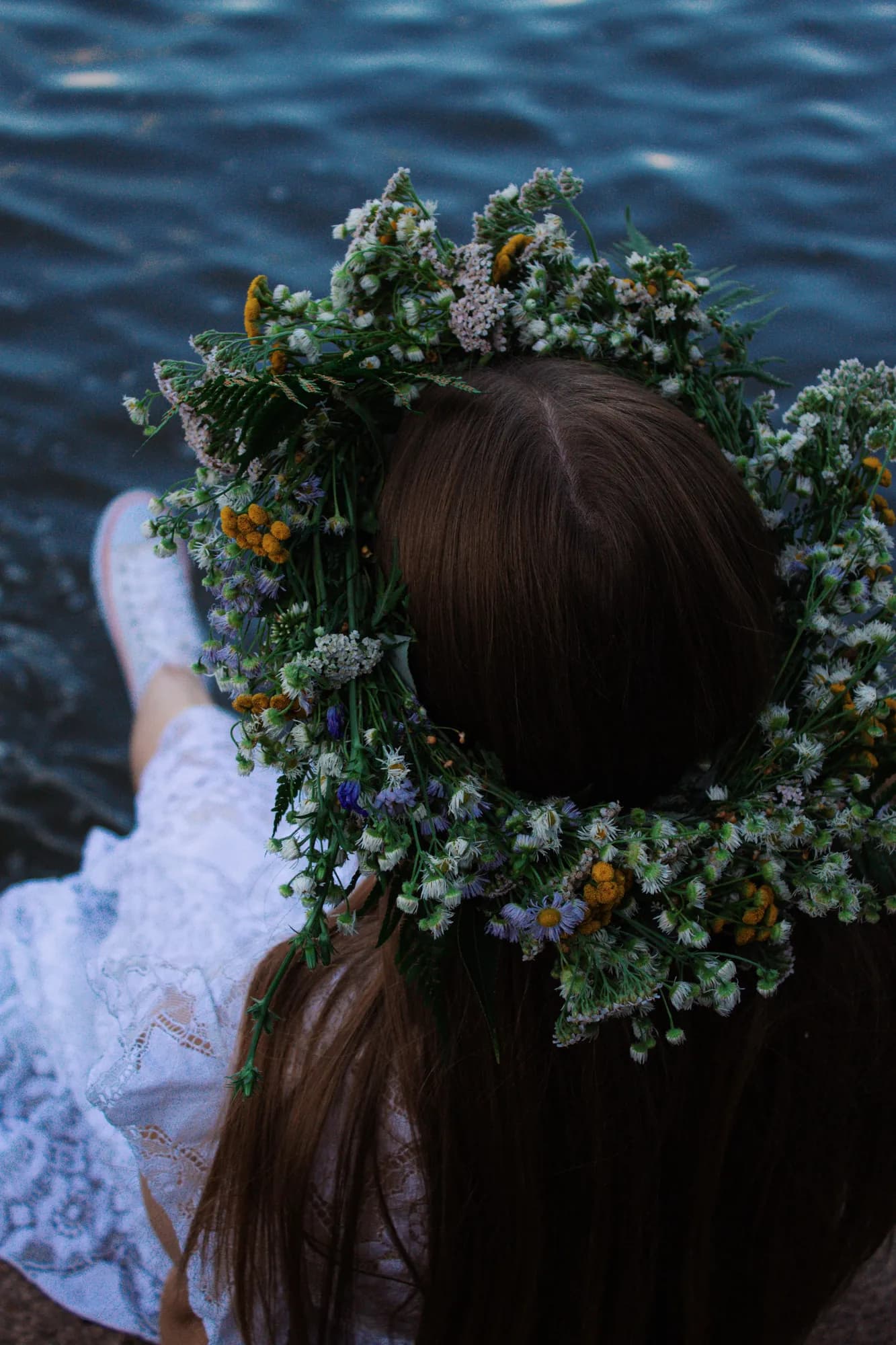 A girl seen from behind wearing a thick crown of white and yellow wildflowers, sitting in a white lace dress beside dark water