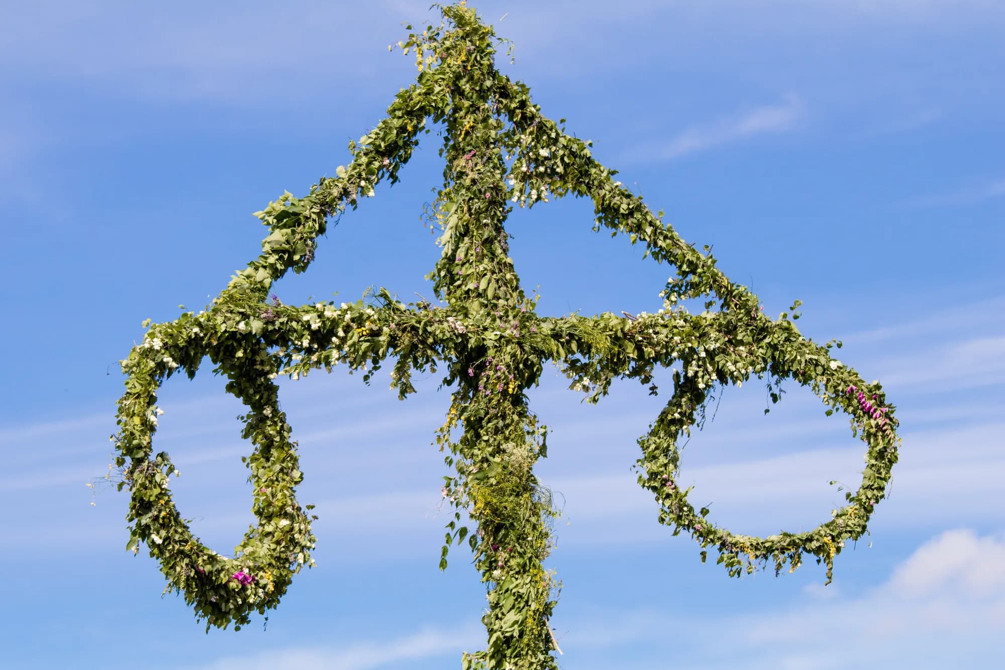 A Swedish Midsummer pole wrapped in green leaves and wildflowers, a cross with two hanging rings, against a blue summer sky