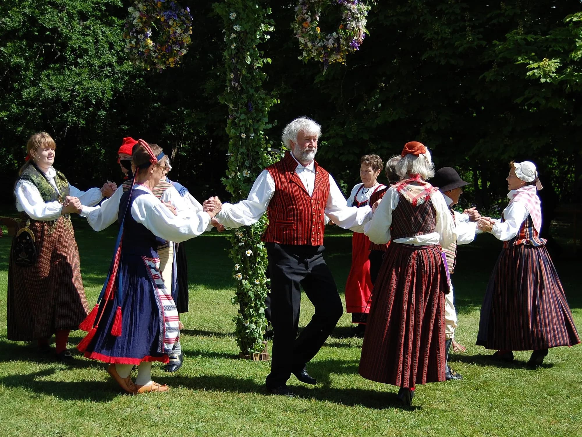 Dancers in traditional folk costume holding hands in a ring on the grass beside a leaf-wrapped pole, flower garlands hanging above