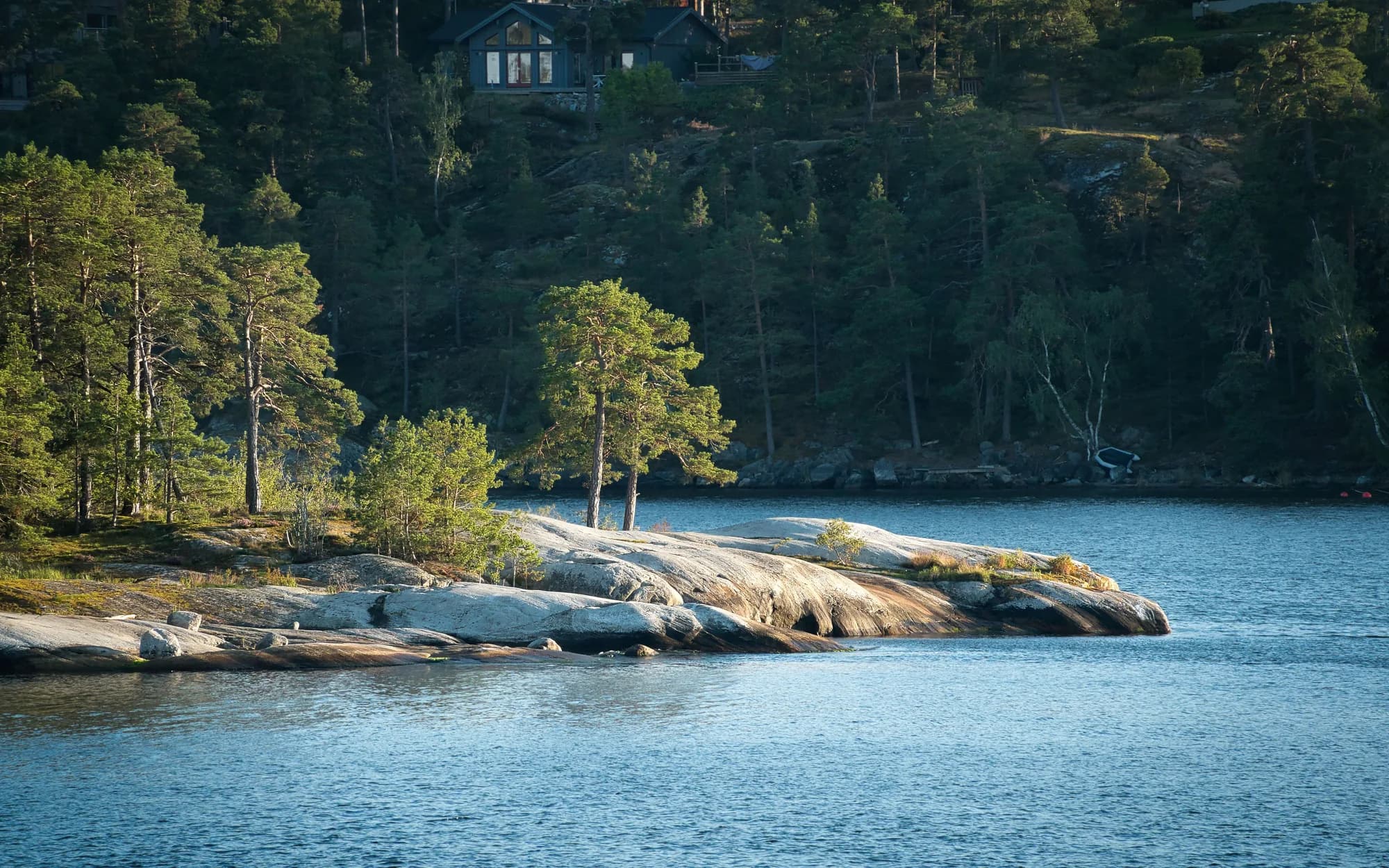 Smooth pale granite running into blue water beneath tall pines, with a wooden house half hidden in the forest above, in low summer evening light