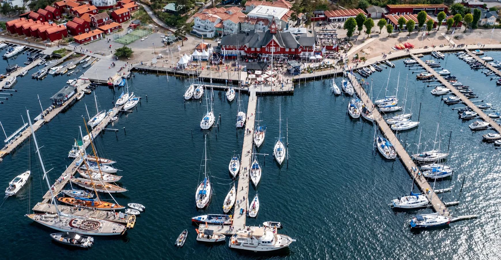 Aerial view of Sandhamn's guest harbour in the Stockholm archipelago, sailing yachts moored along long wooden piers below the red Seglarhotell and red-roofed houses