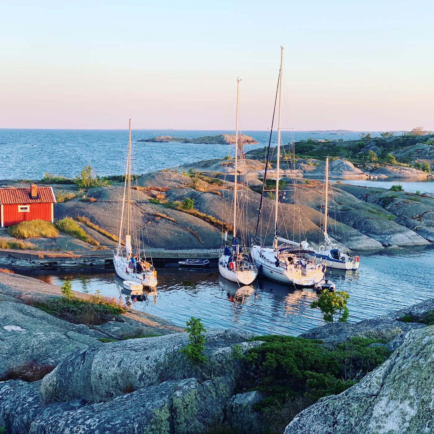 Four sailing yachts moored in a small natural harbour between smooth granite rocks, a red wooden cottage on the shore and the open sea beyond at dusk